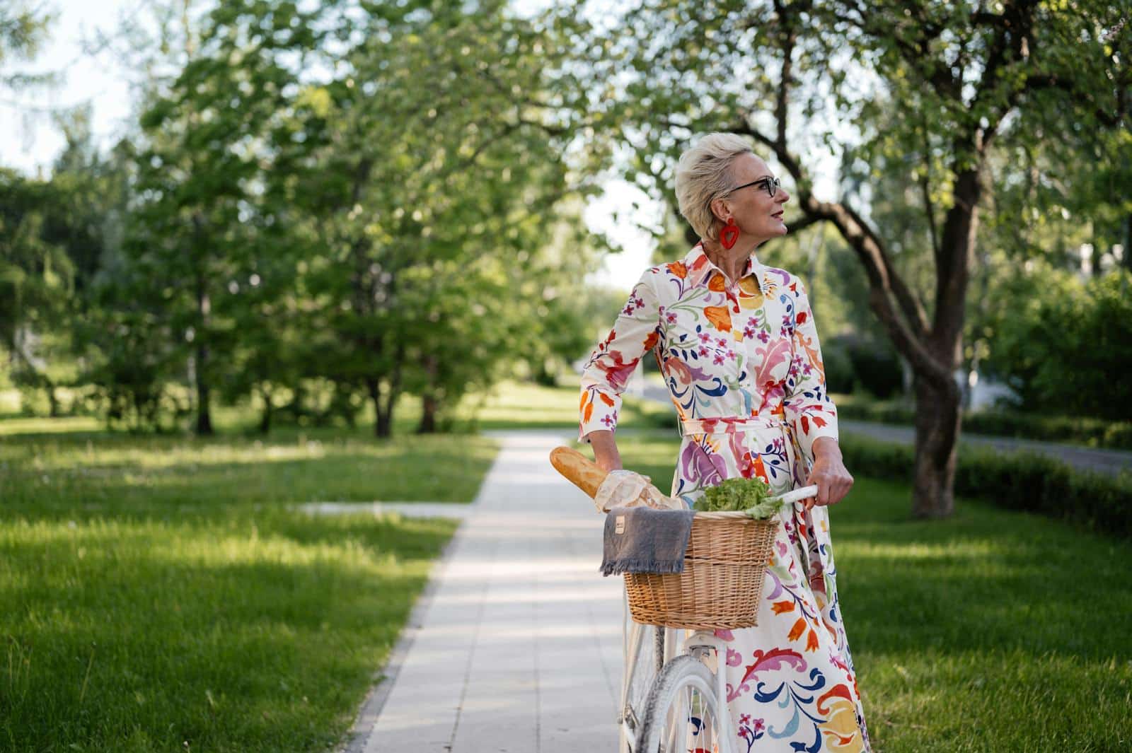 Fashionably dressed woman rides a bicycle in a lush park setting with a basket of groceries.